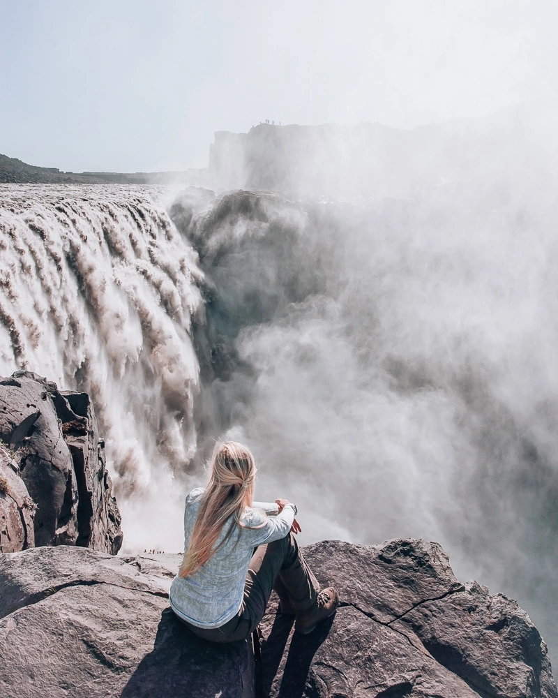 Woman looking at Dettifoss waterfall in Iceland from the edge of the cliff into the canyon. Find the top 5 waterfalls in Iceland that you don't want to miss on a Ring Road road trip. 