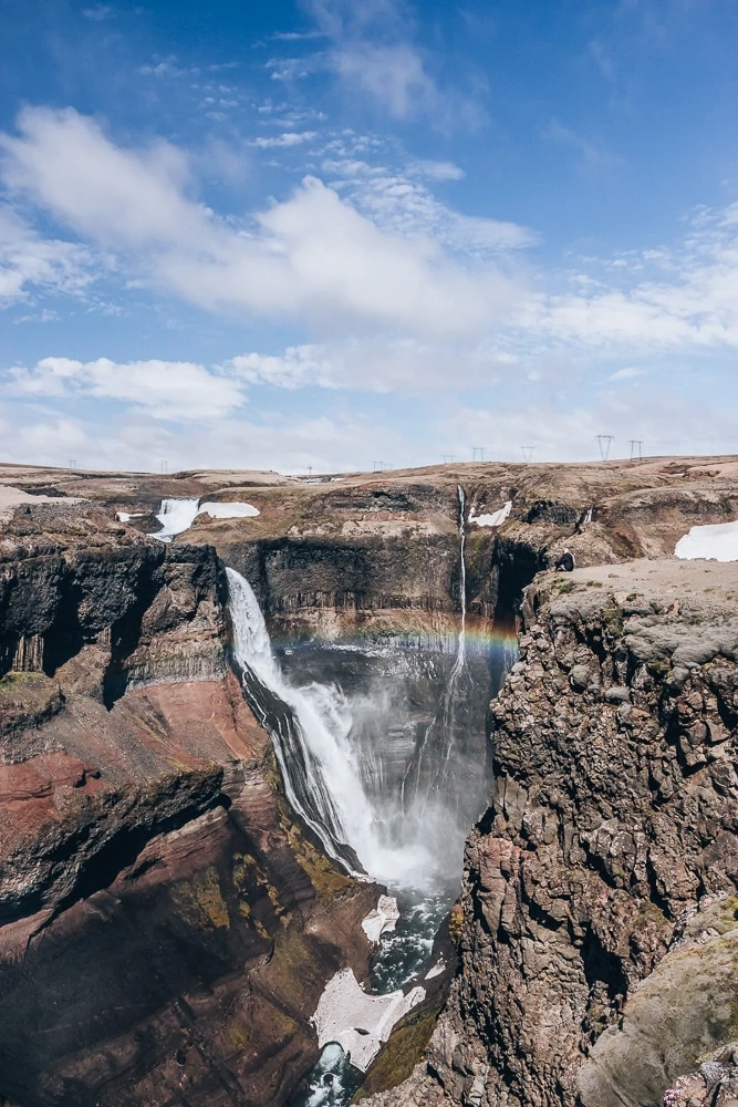 Woman sitting on the edge of a cliff looking at Granni waterfall with a rainbow in southeast Iceland. Find the top 5 waterfalls in Iceland that you don't want to miss on a Ring Road road trip. 
