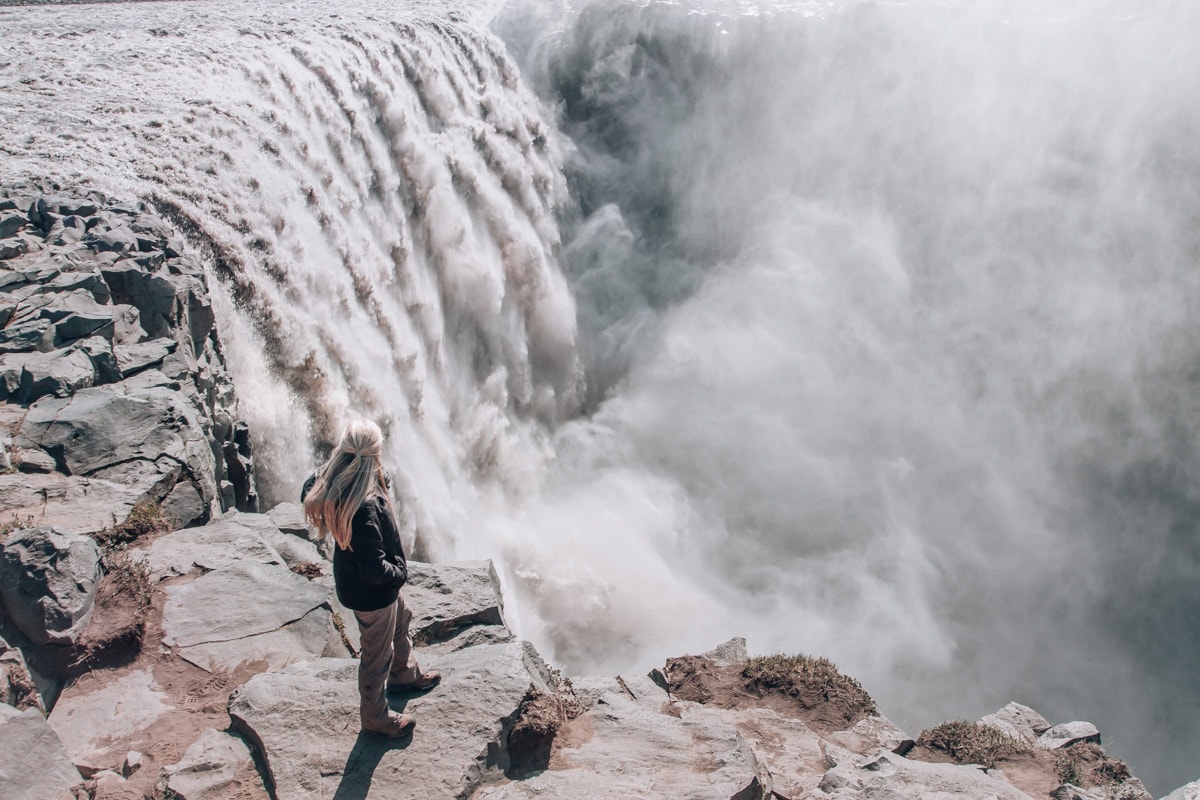 Woman standing on the edge of Dettifoss Waterfall in Iceland. Get all the best stops on an Iceland road trip with our 6 day Ring Road itinerary