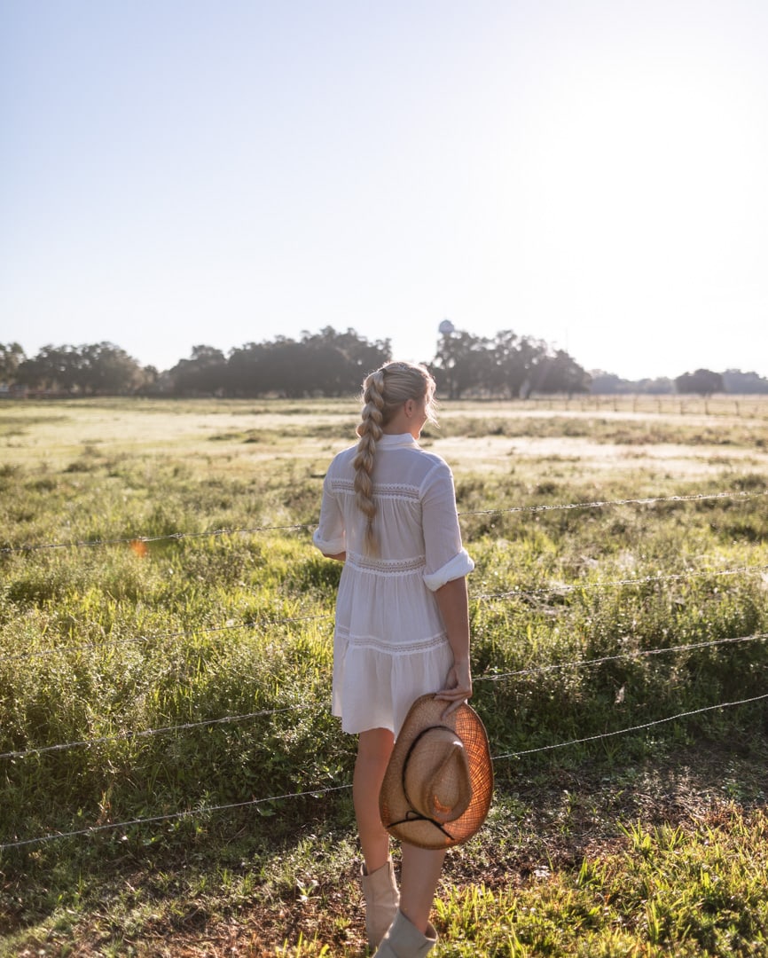 Woman in white dress from Aggie Swimwear, cowboy hat, and boots looking out over pastures while glamping in Florida at Westgate River Ranch.